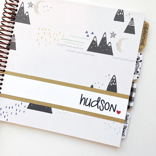 Mountain themed baby book. Light grey cover with black mountain groupings, stars and moons. Personalized with baby's name on the front cover.